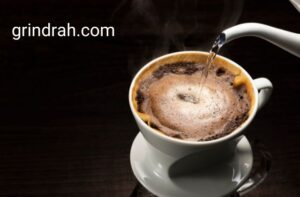 A picture of a brewed coffee in a white mug kept on a small tray and a kettle of hot water pouring on it. The picture has the name of our website grindrah.com tagged on it.